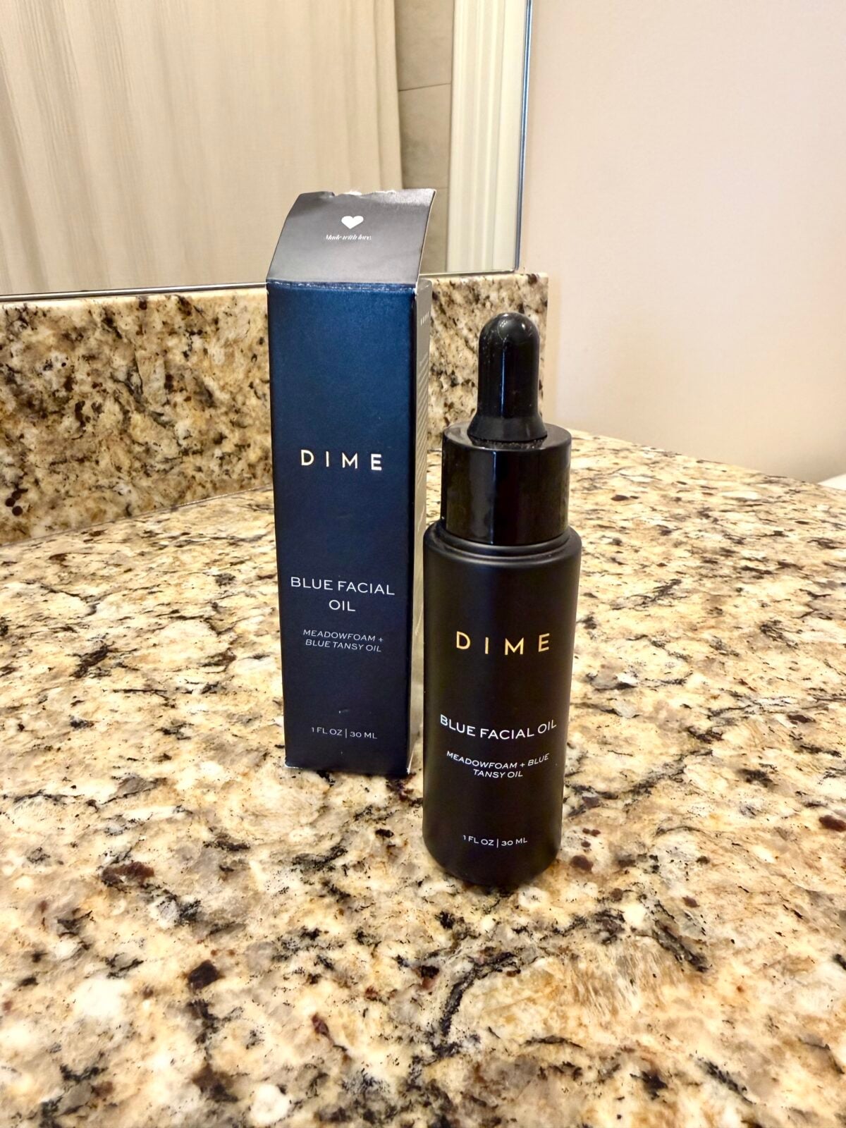 A bottle of DIME Blue Facial Oil with a dropper is placed next to its matching box on a granite bathroom countertop, with a mirror and beige wall in the background.