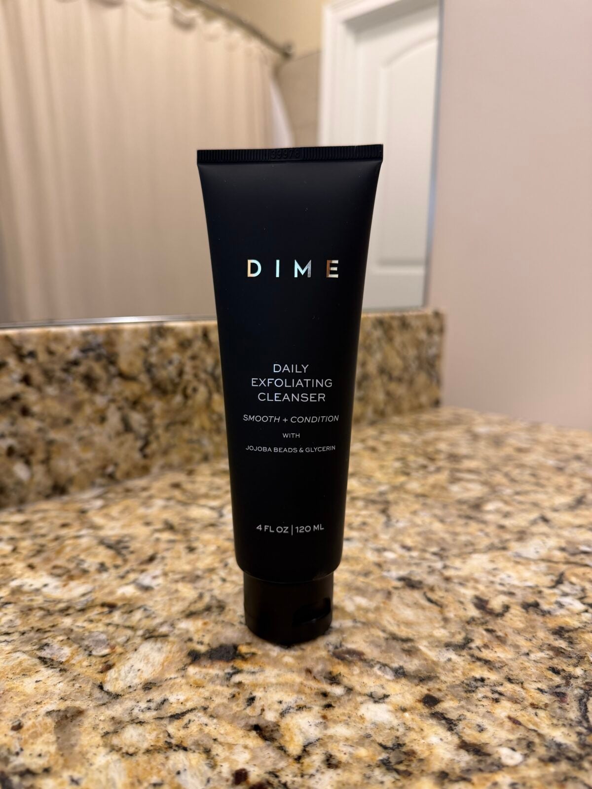 A black tube labeled DIME Daily Exfoliating Cleanser stands upright on a granite bathroom countertop. The background shows a shower curtain and a white door.