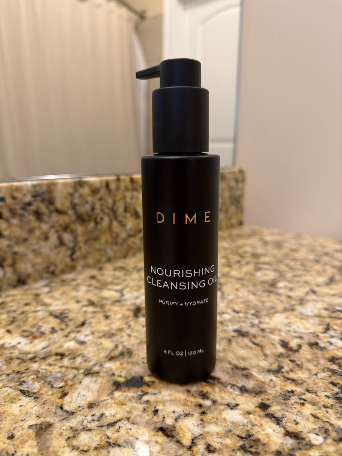 A black bottle of DIME Nourishing Cleansing Oil sits on a granite bathroom counter, with a shower curtain and white door visible in the background.