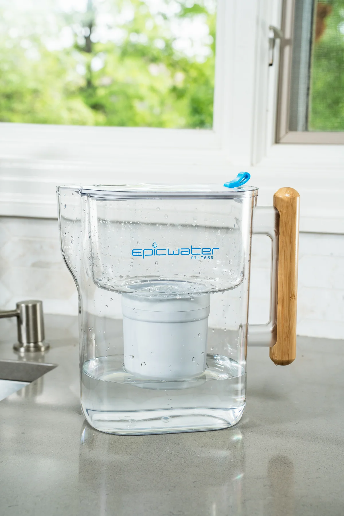The photo shows an Epic Water Filters pitcher with a bamboo handle and blue accents, sitting on a kitchen countertop in front of a window.