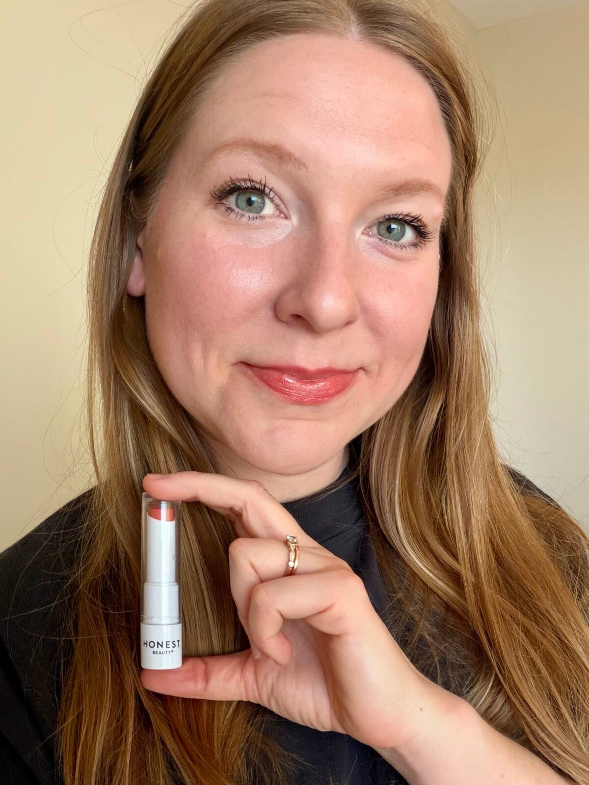 Woman holding an Honest Beauty tinted lip balm in a coral shade, wearing the product on her lips