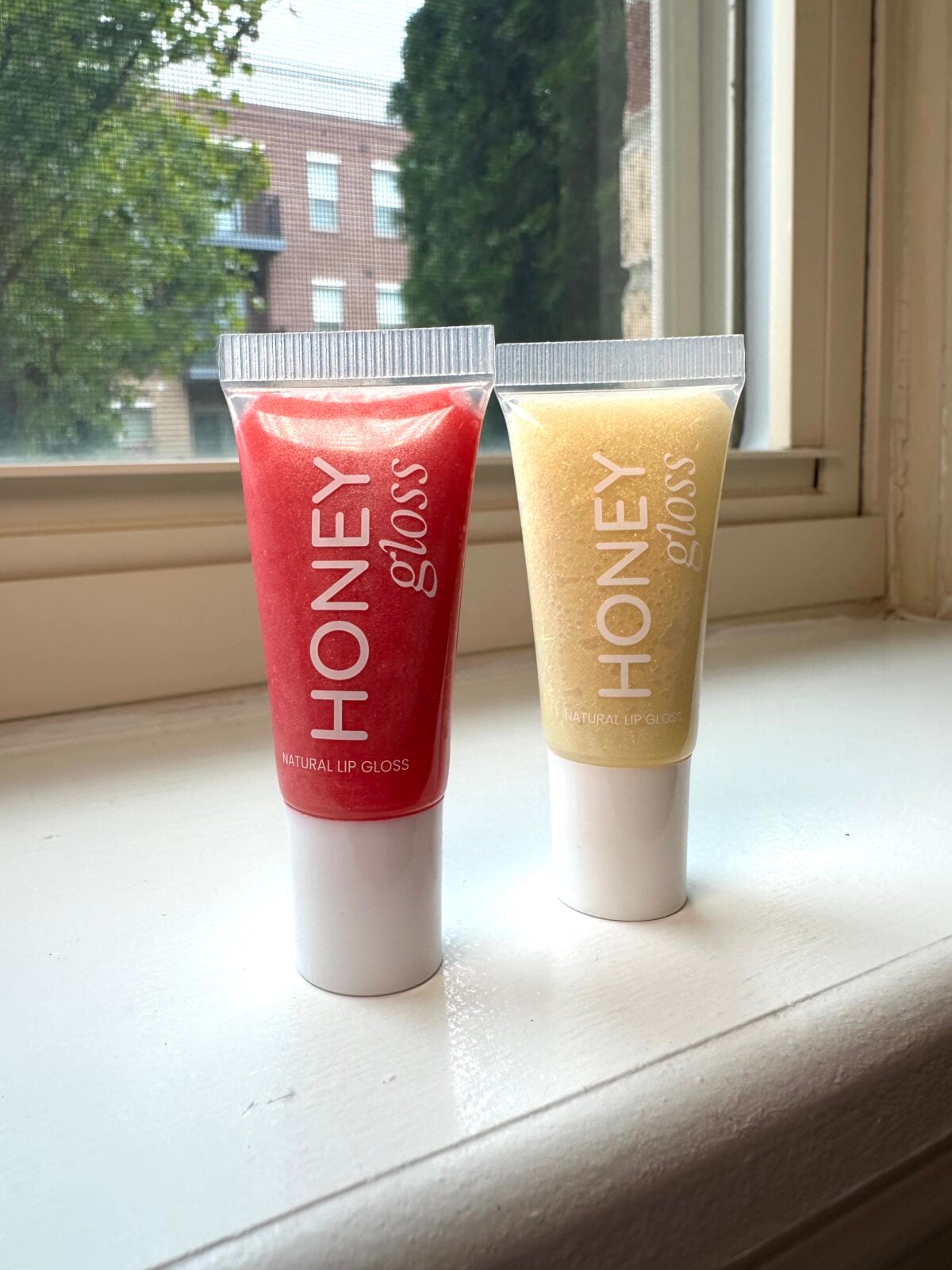 Two tubes of Honey Gloss natural lip gloss in red and gold shimmer shades sitting on a windowsill with natural light