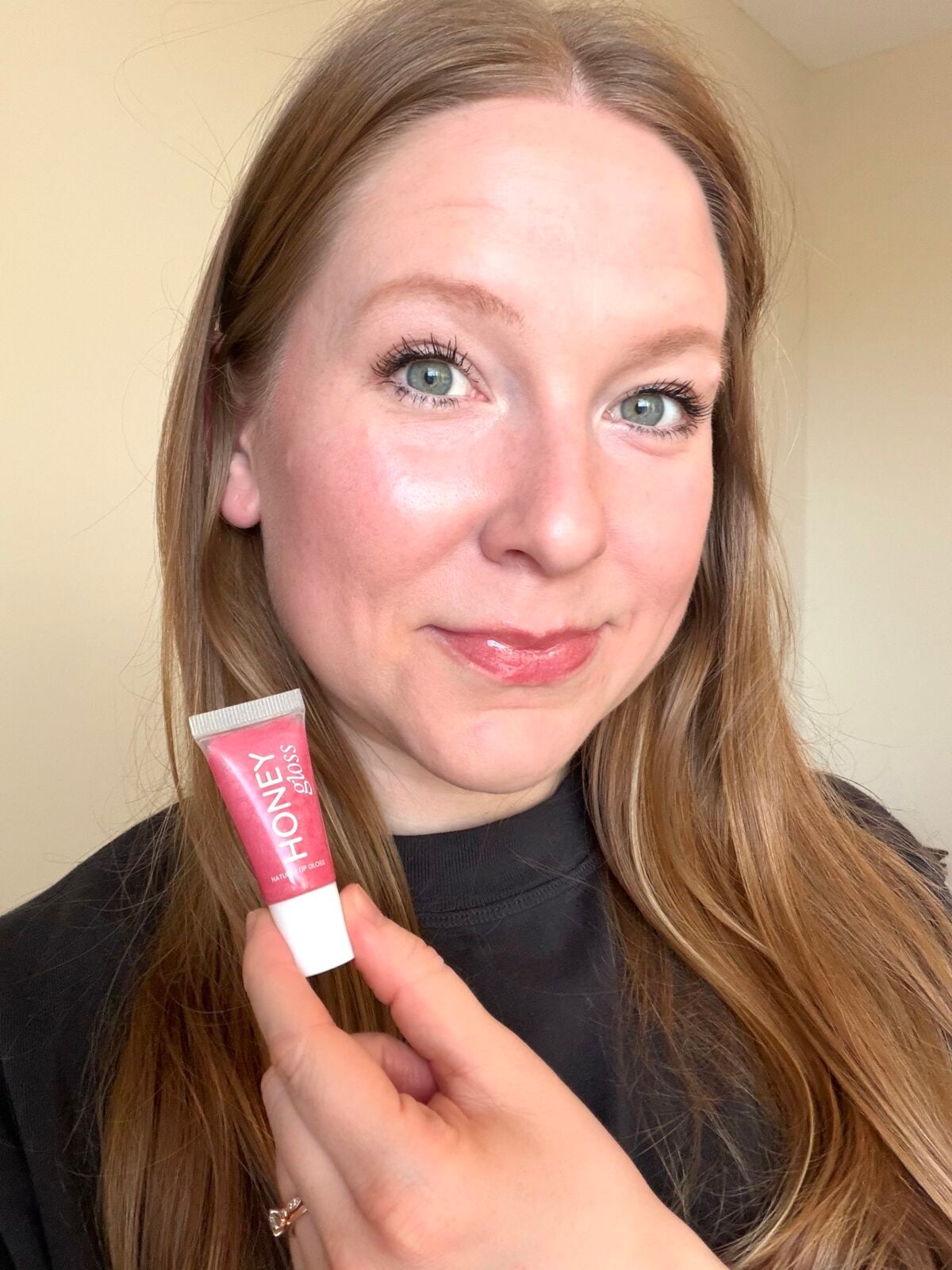 Woman holding a pink tube of Honey Gloss natural lip gloss, wearing the product on her lips