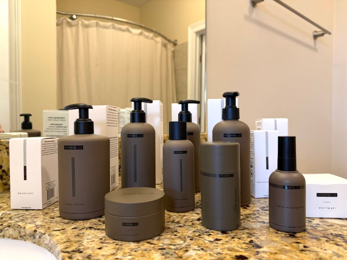 The photo shows an assortment of matte-brown personal care bottles and jars from the brand Hygiene Lab, including body lotion, face wash, and deodorant, arranged on a granite bathroom countertop.