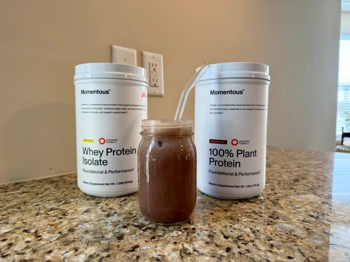 Two white containers of Momentous protein powder, one labeled &ldquo;Whey Protein Isolate&rdquo; and the other &ldquo;100% Plant Protein,&rdquo; are on a kitchen counter behind a glass jar filled with chocolate-colored liquid and a clear straw.