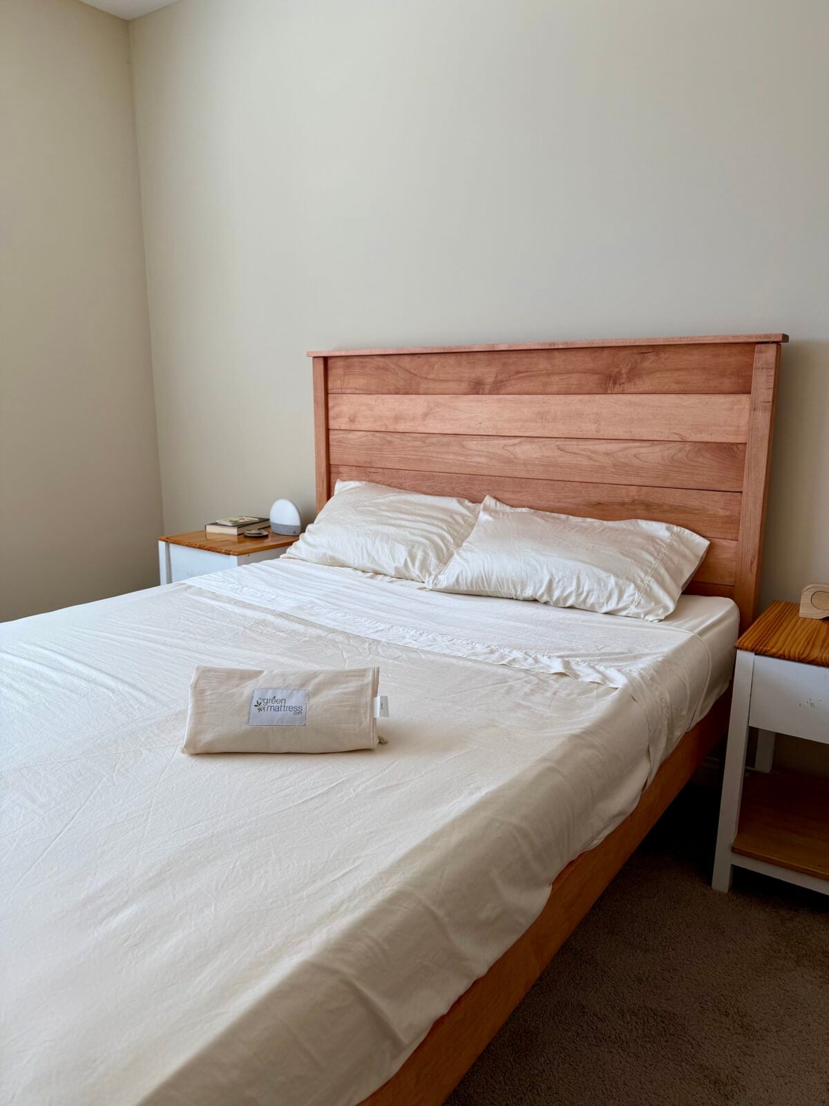 A neatly made bed with white sheets and two pillows sits against a beige wall. A wooden headboard and two small bedside tables, one with stacked books and a lamp, complete the scene. A folded towel lies on the bed.