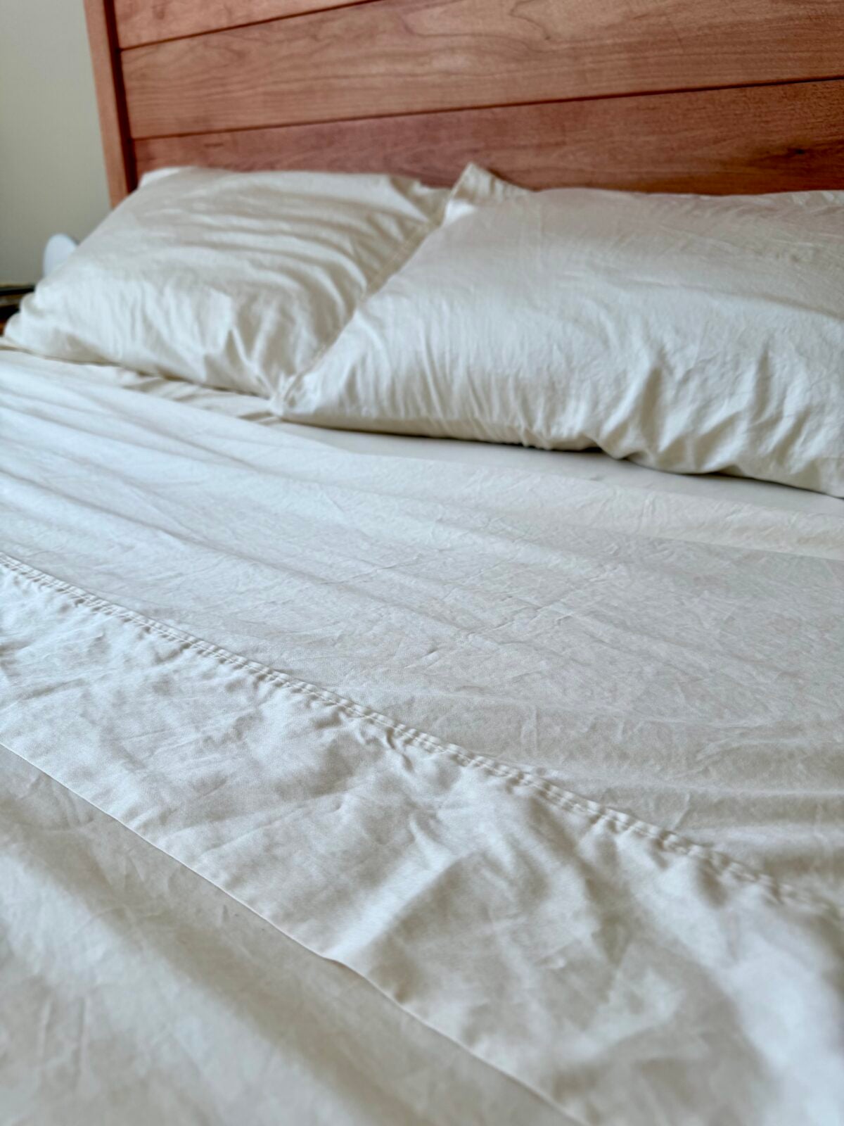 A neatly made bed with off-white sheets and two pillows against a wooden headboard. The bedding appears smooth and clean, with slight creases visible in the fabric.