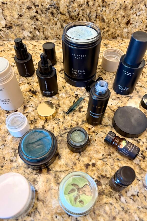 A variety of skincare products, including jars, bottles, and tubs with mostly black packaging, are arranged on a speckled granite countertop. Some products have blue or green creams visible inside open containers.