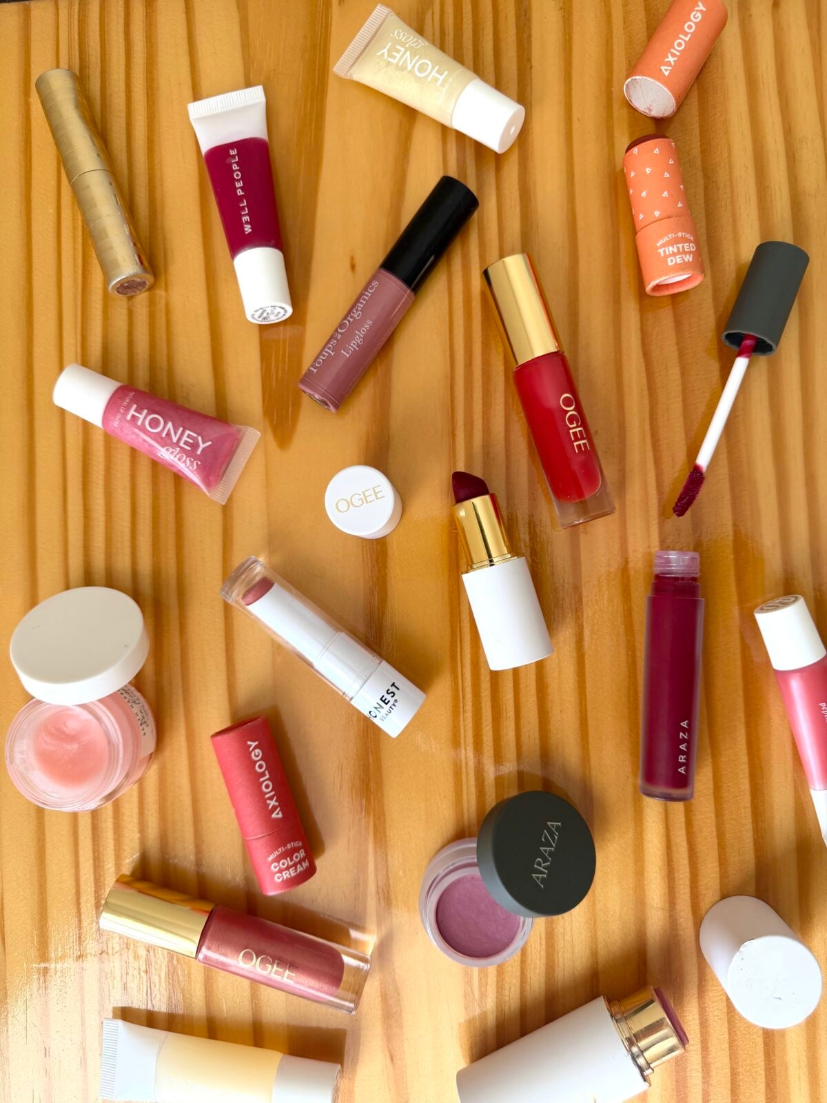 Non-toxic lipstick and lip gloss products scattered on a wooden surface, including brands Ogee, Axiology, Araza, Well People, Honest Beauty, and Toups & Co Organics in various shades of pink, red, and berry.