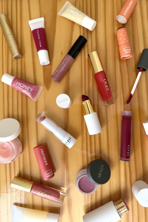 Non-toxic lipstick and lip gloss products scattered on a wooden surface, including brands Ogee, Axiology, Araza, Well People, Honest Beauty, and Toups & Co Organics in various shades of pink, red, and berry.