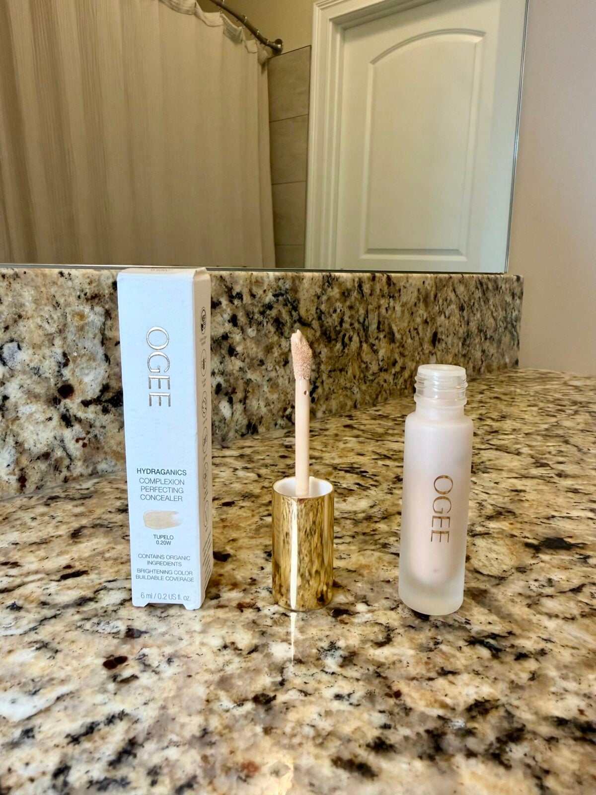 A tube of OGEE hydragamics sculpted complexion concealer with a gold-capped applicator is open and standing next to its white box on a speckled bathroom countertop. A mirror and shower curtain are in the background.