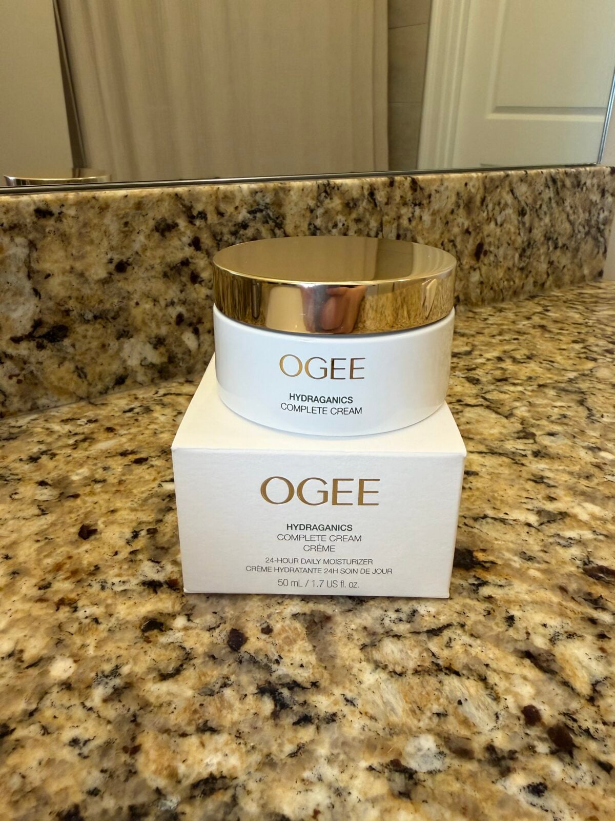 A jar of Ogee Hydraganics Complete Cream sits on top of its matching product box on a granite bathroom countertop, with a mirror and part of a door visible in the background.