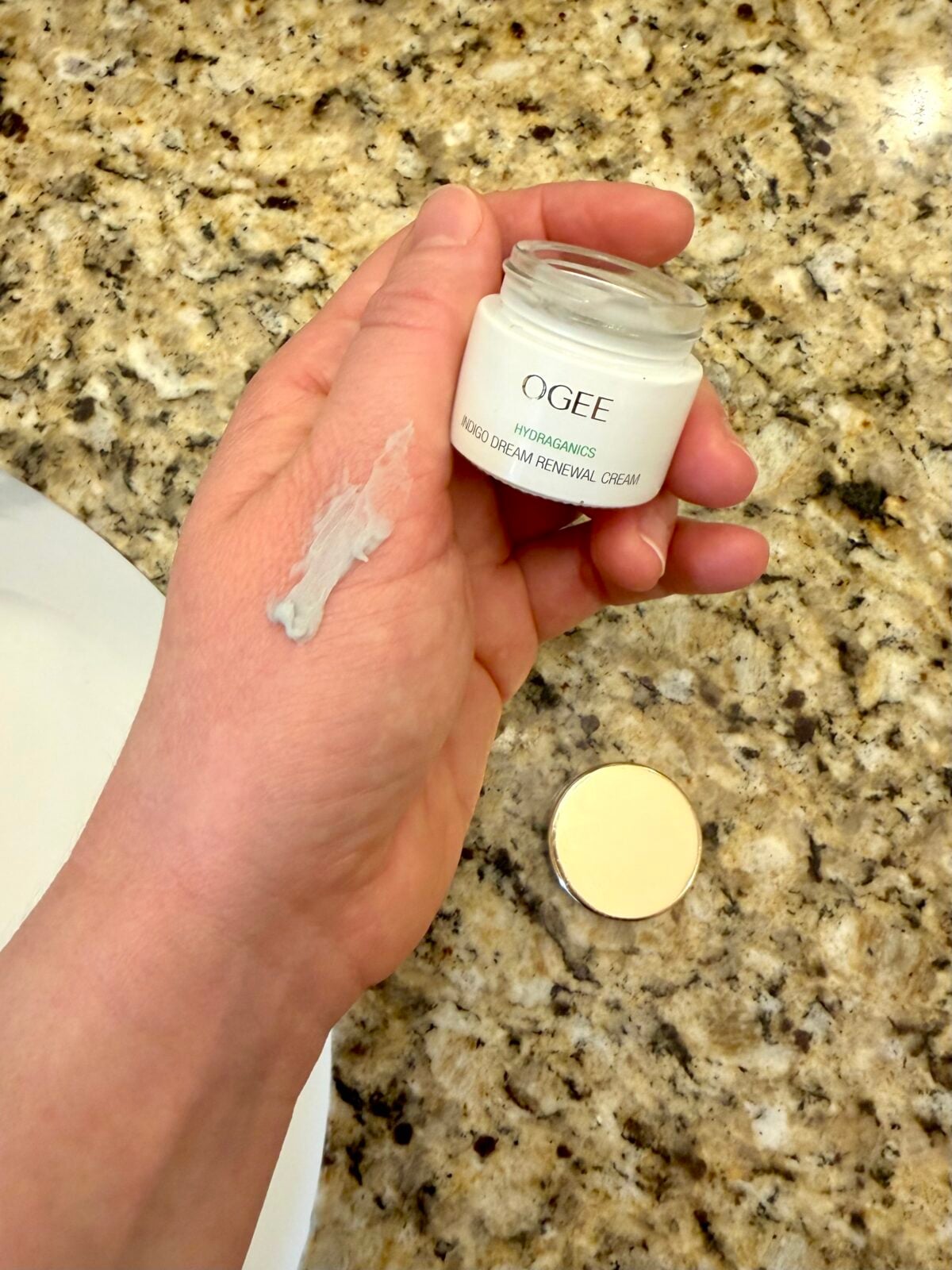 A hand holds an open jar of Ogee Hydroganics Rose Dream Renewal Cream with a dab of the cream on the back of the hand. The lid is placed on a granite countertop nearby.