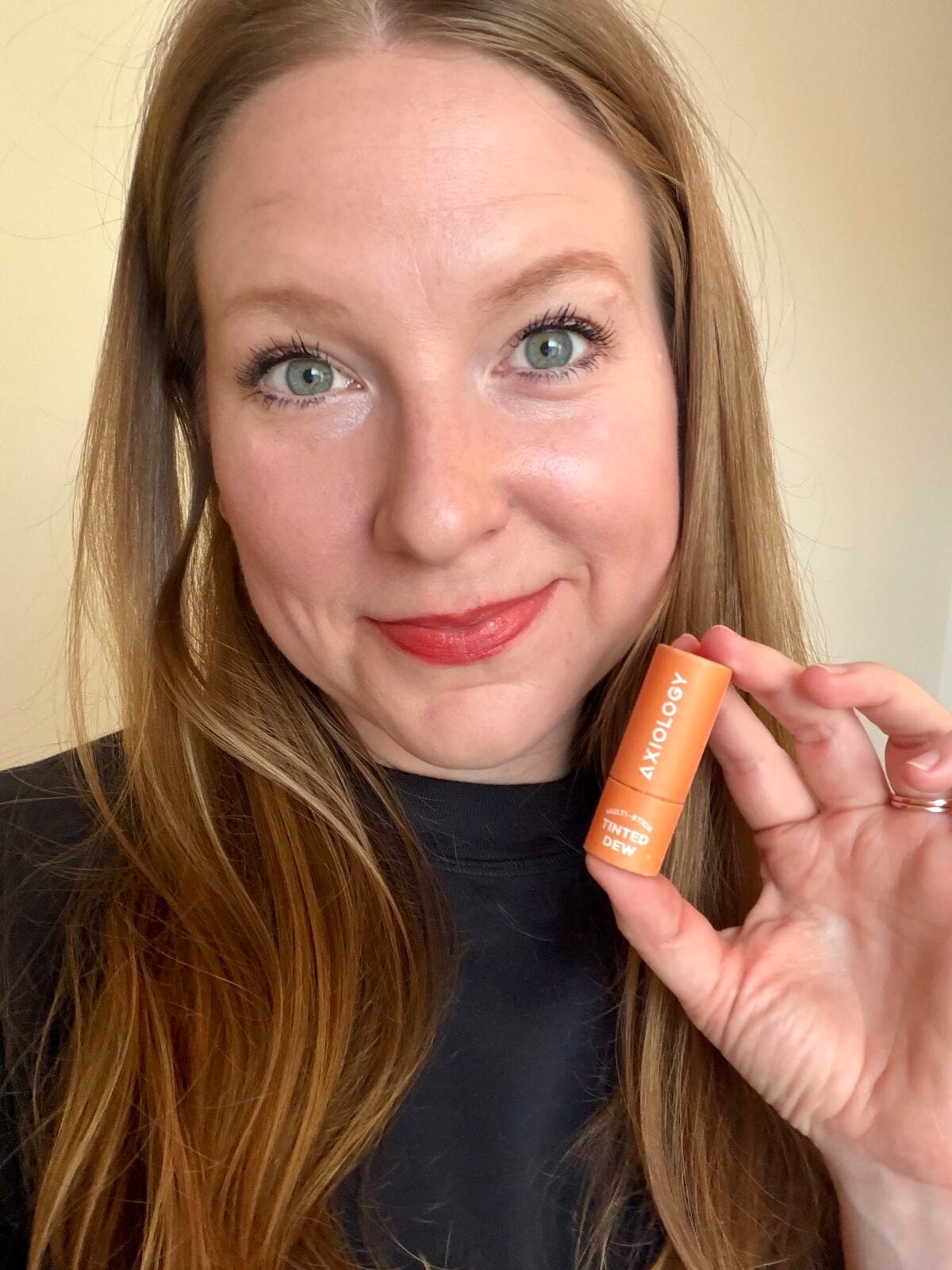 Woman holding a small orange Axiology Multi-Stick Tinted Dew in plastic-free packaging, wearing a red-tinted lip color