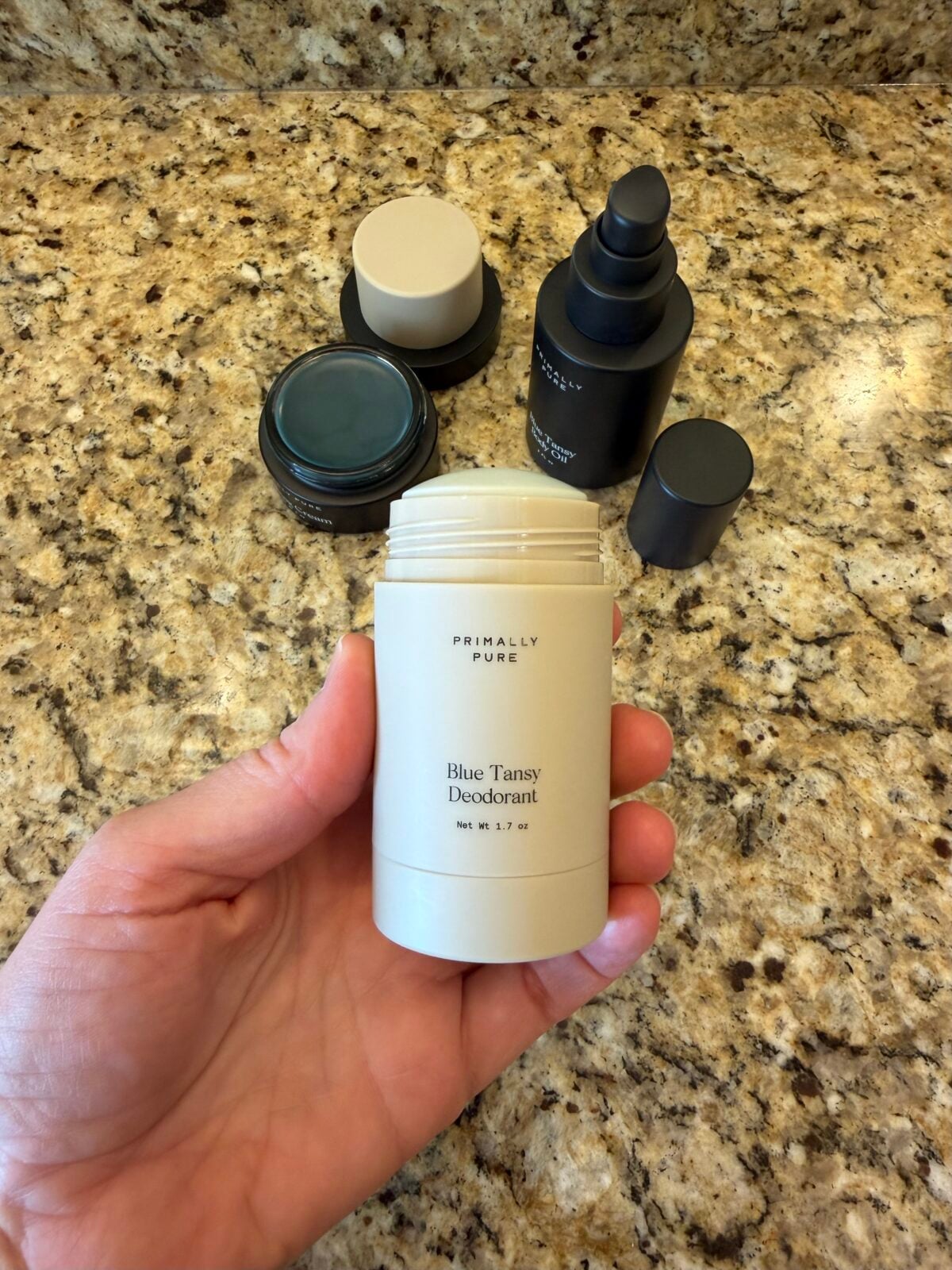 A hand holds an open tube of Primally Pure Blue Tansy Deodorant above a granite countertop, with three closed skincare containers in the background.