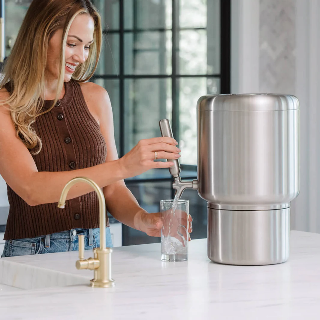 A woman in a brown ribbed top is shown smiling while dispensing water into a glass from a large, cylindrical stainless steel filtration system on a white kitchen island.