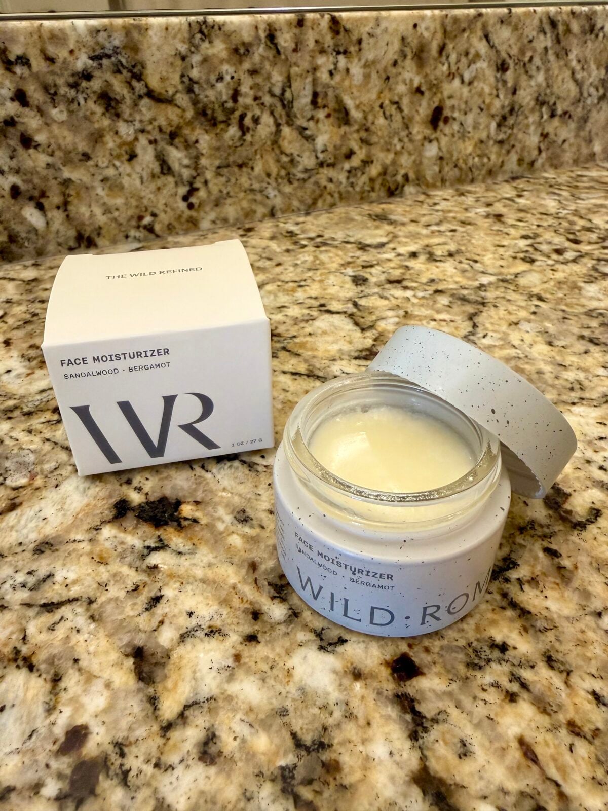 A jar of Wild Rose face moisturizer with the lid off sits on a granite countertop next to its white product box labeled Face Moisturizer Sandalwood • Bergamot.