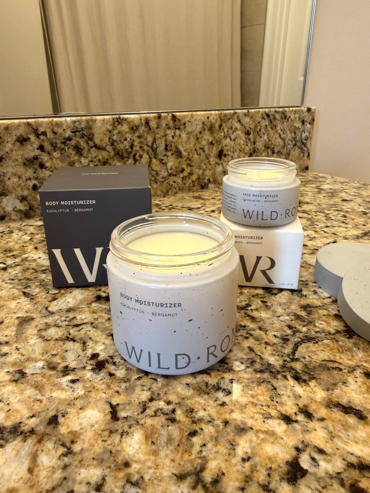 An open jar of Wild Roar body moisturizer sits on a granite bathroom countertop next to its box and a second, closed jar of the same product. The label mentions eucalyptus and bergamot.