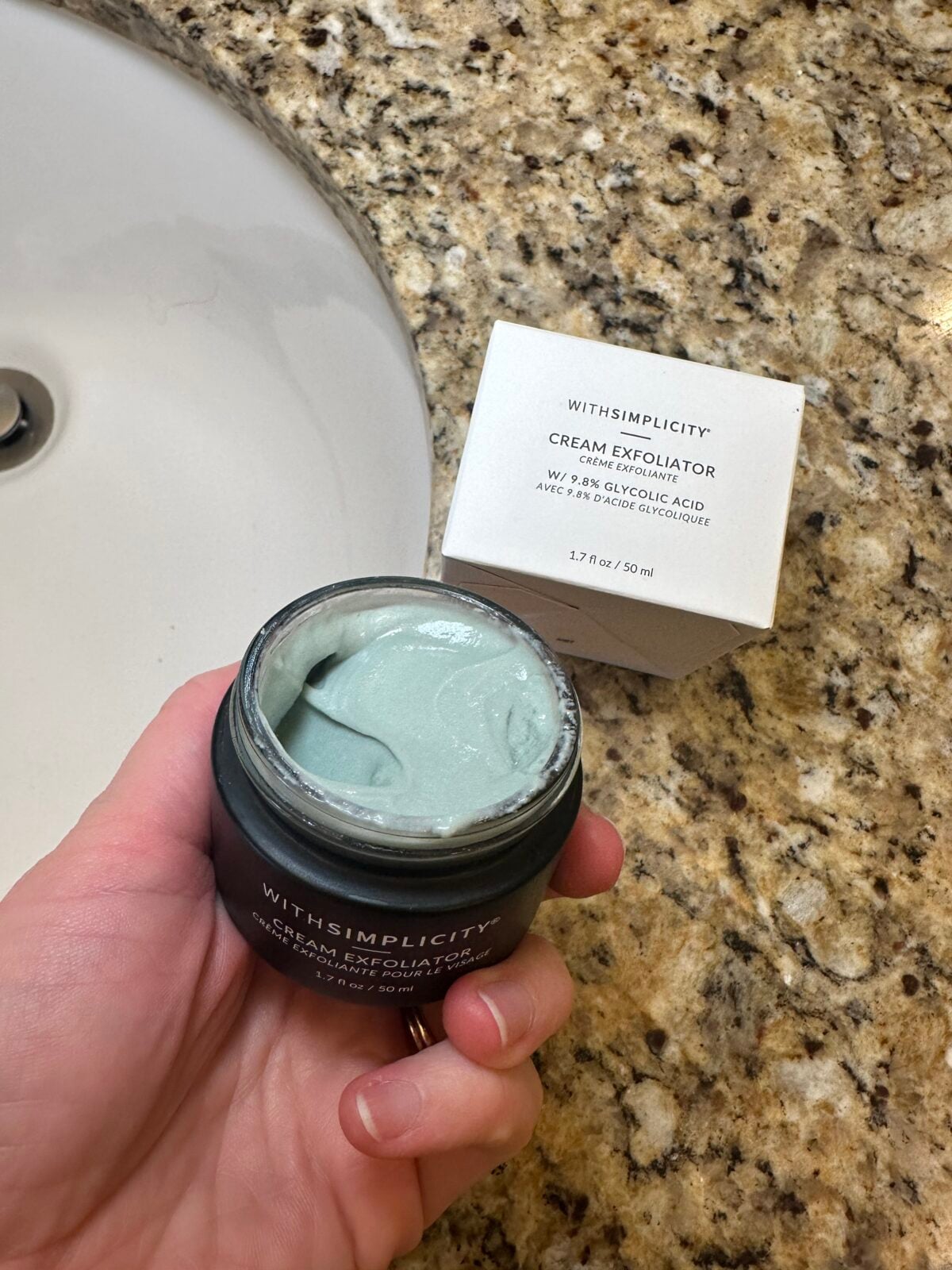 A hand holds an open jar of WITHSIMPLICITY cream exfoliator showing pale green product inside, with the product&rsquo;s white box in the background on a beige granite bathroom counter next to a sink.