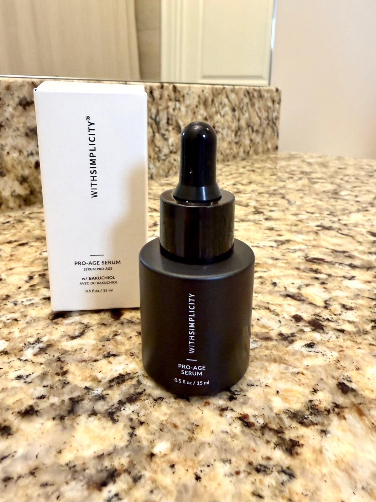 A black dropper bottle labeled WITHSIMPLICITY PRO-AGE SERUM stands on a granite countertop, with its white product box positioned behind it. The bottle contains 0.5 fl oz (15 ml) of serum.
