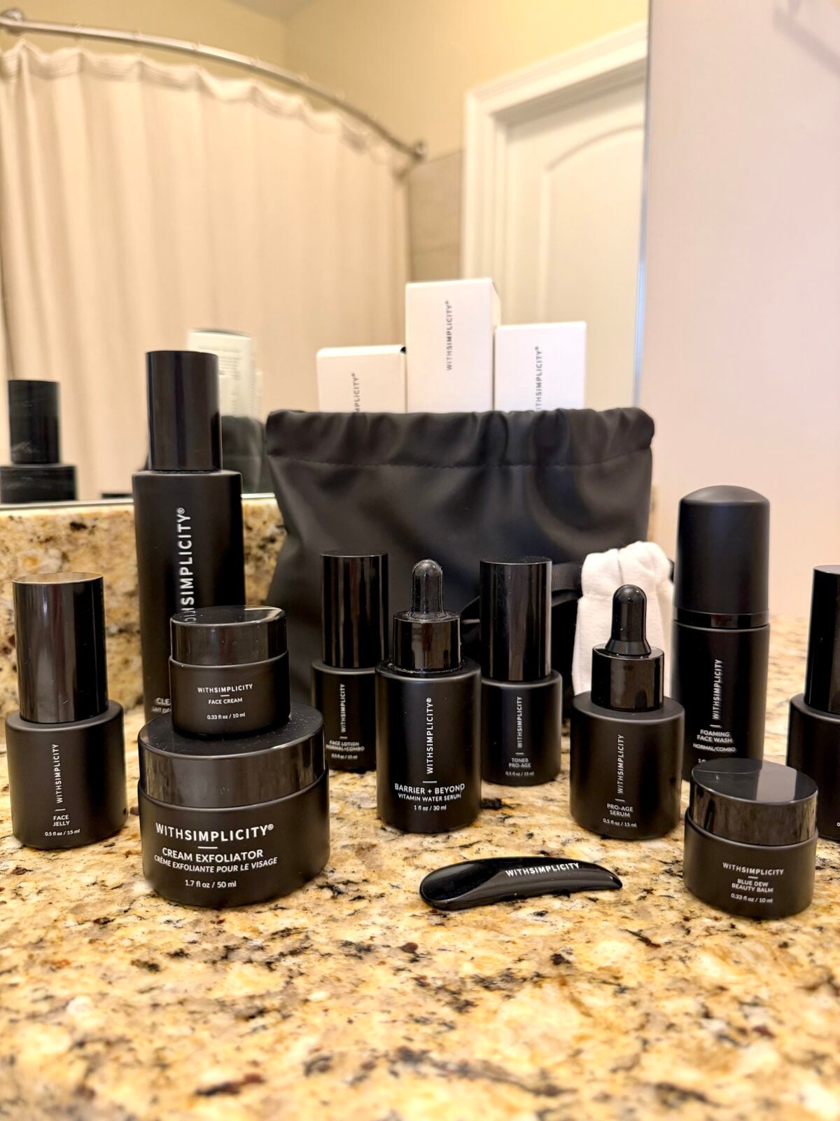 A collection of black skincare product bottles and jars labeled WithSimplicity arranged on a granite bathroom countertop, with a black toiletry bag and boxed products in the background.