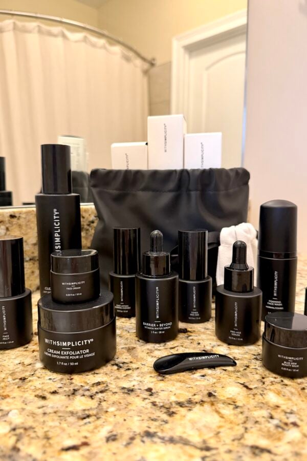 A collection of black skincare product bottles and jars labeled WithSimplicity arranged on a granite bathroom countertop, with a black toiletry bag and boxed products in the background.