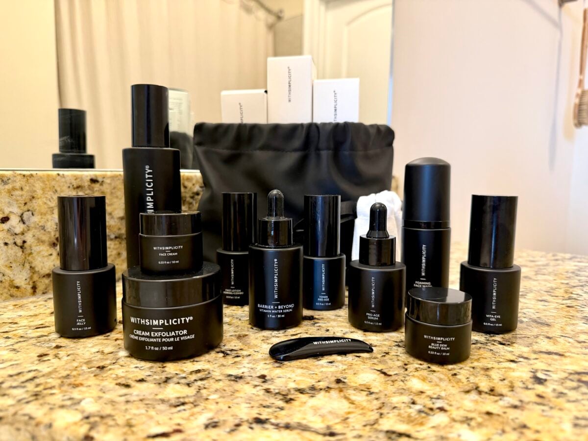 A collection of black skincare product containers labeled “WITHSIMPLICITY” arranged on a bathroom countertop, with a black cosmetic bag and boxes in the background. The products include jars, bottles, and droppers.