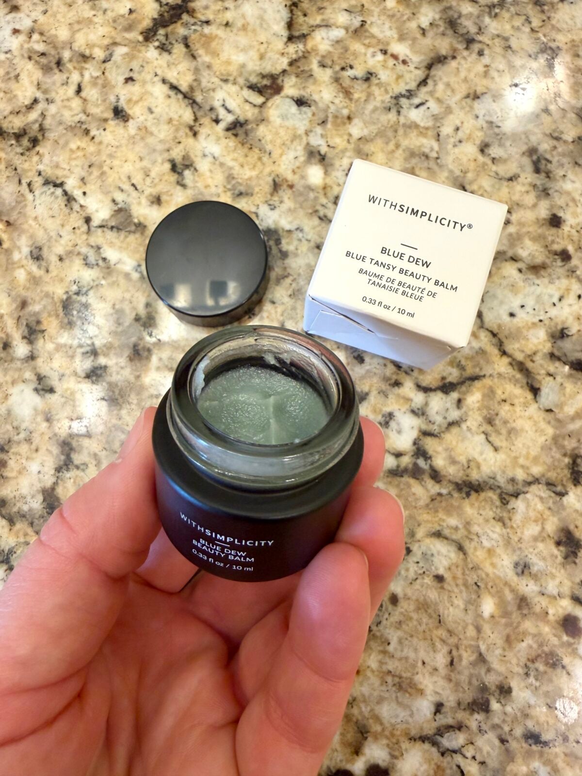 A hand holds an open jar of blue beauty balm labeled &ldquo;WITHSIMPLICITY BLUE DEW,&rdquo; with the lid resting nearby and the product&rsquo;s white box sitting on a speckled granite countertop.