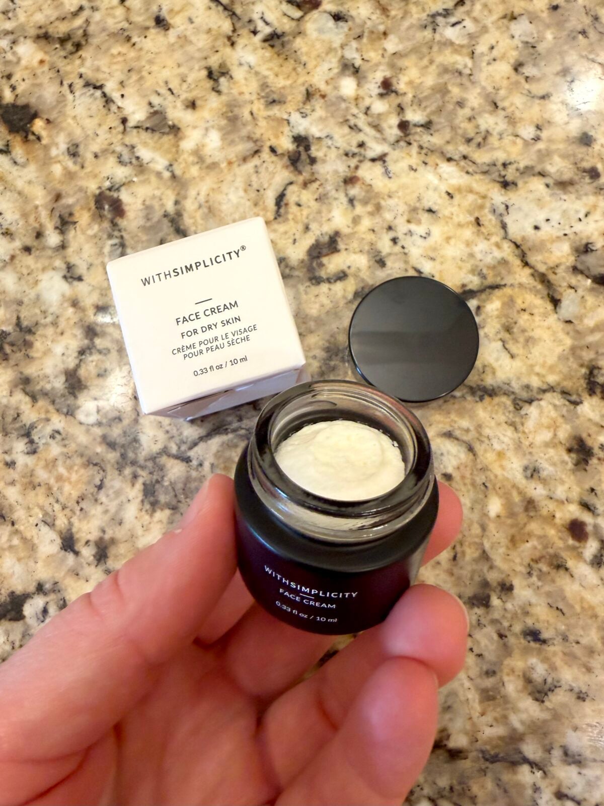 A hand holds an open jar of withSimplicity face cream for dry skin; the jar and its packaging rest on a speckled granite countertop. The jars lid is next to the container.