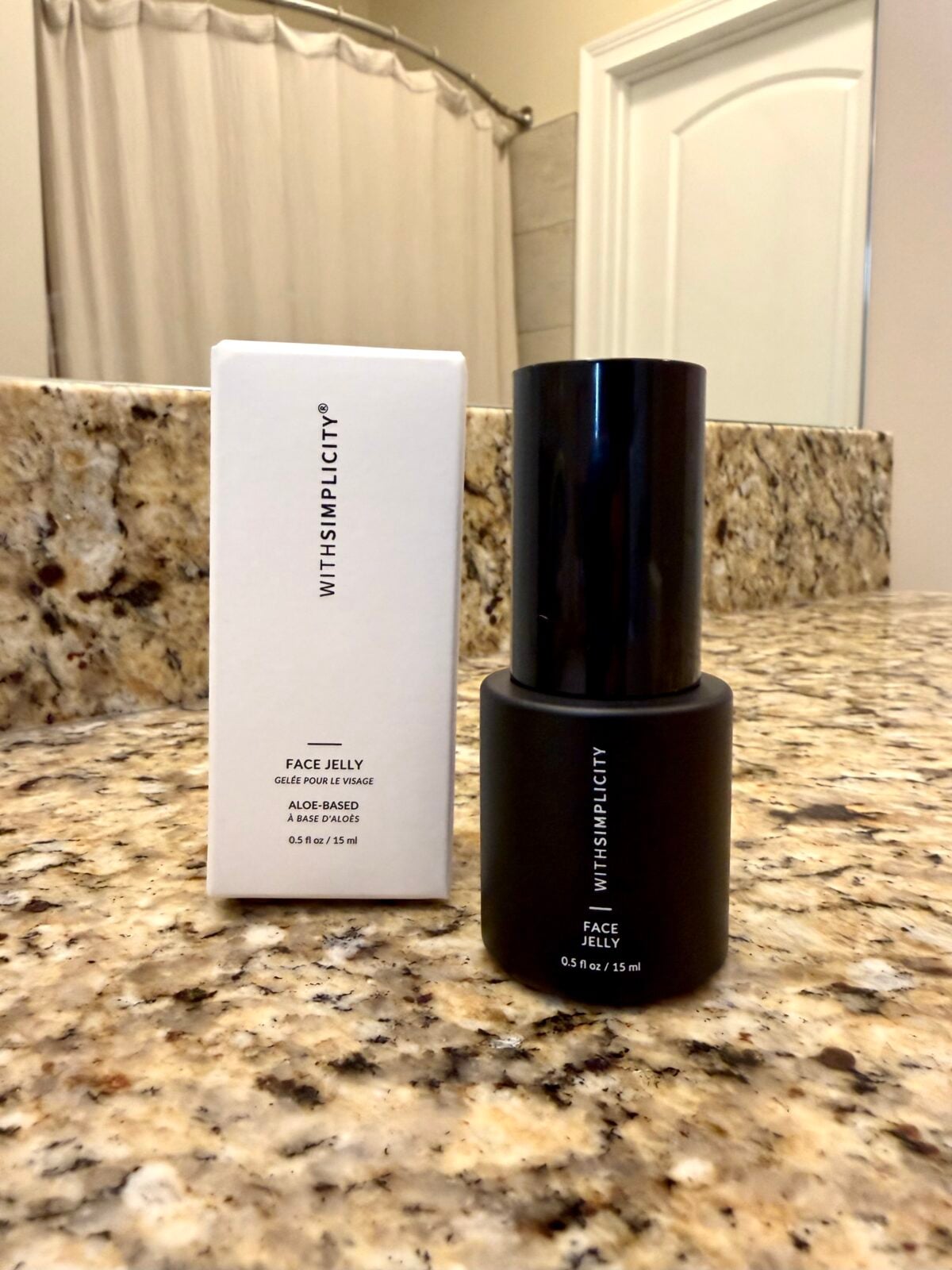 A black bottle labeled &ldquo;Face Jelly&rdquo; and a white rectangular box with &ldquo;WITH SIMPLICITY&rdquo; branding sit on a granite bathroom counter with a shower curtain and white door visible in the background.