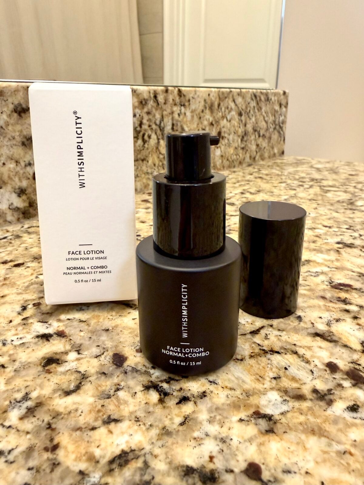 A black bottle of WITHSIMPLICITY face lotion with a pump and black cap sits on a bathroom counter next to its white product box. Both display product details for normal and combination skin.