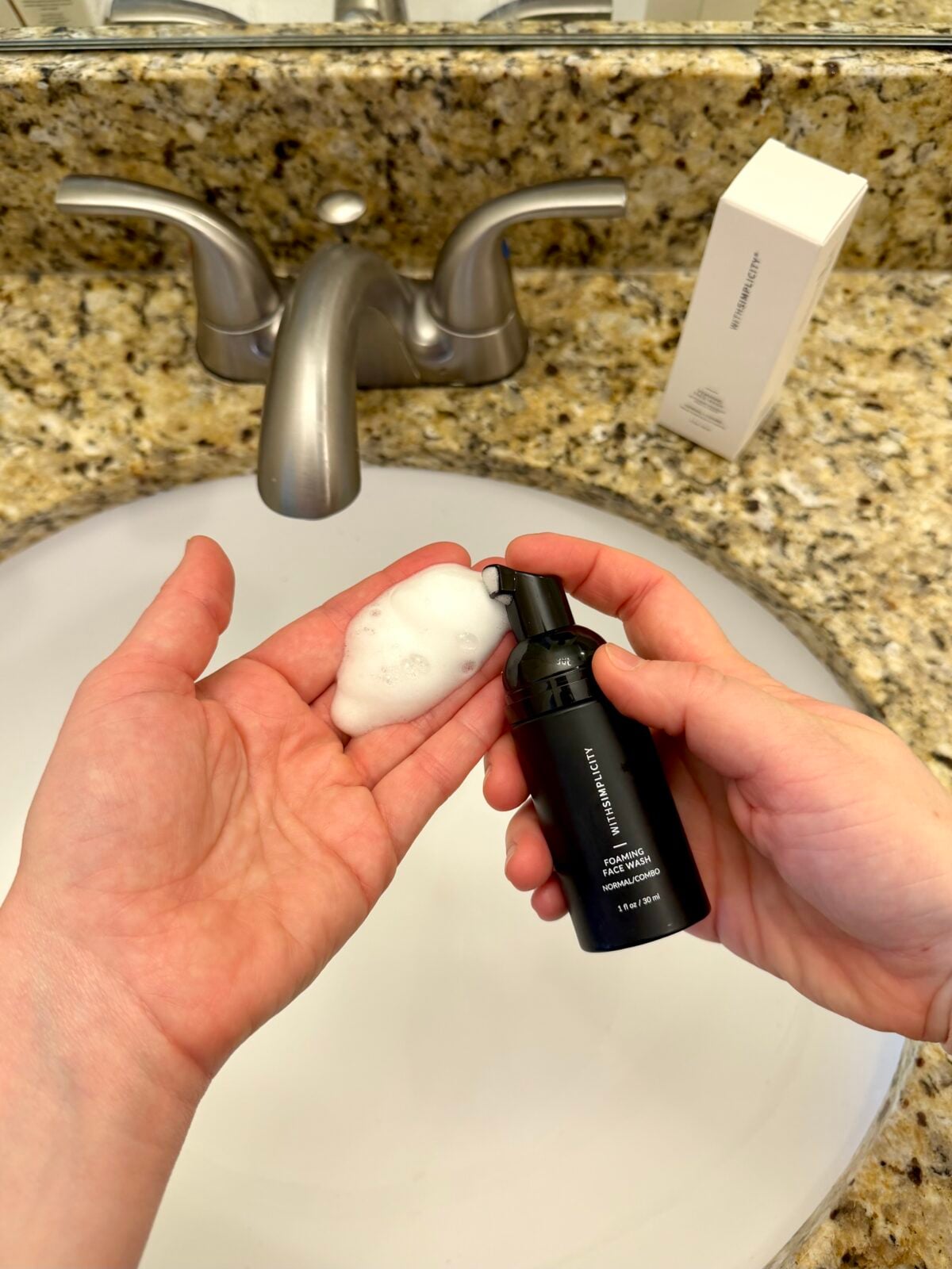 A person dispenses foaming face cleanser from a black bottle onto their palm over a bathroom sink with a granite countertop; the product box is visible near the faucet.