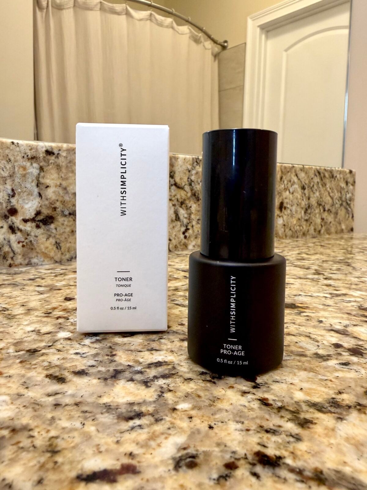 A black bottle labeled WITH SIMPLICITY TONER PRO-AGE 0.5 fl oz / 15ml is placed on a granite countertop next to its matching white packaging box in a bathroom setting.