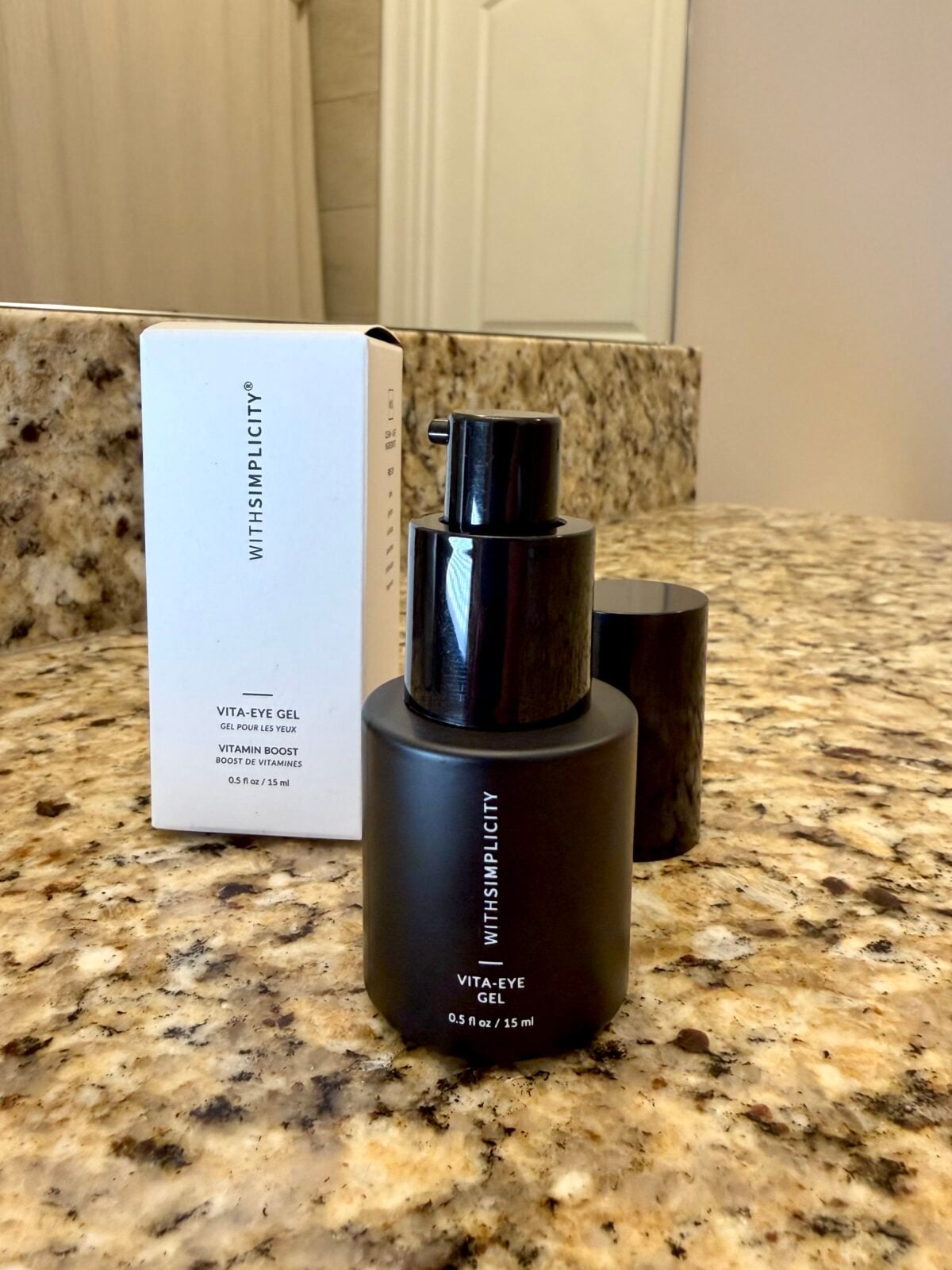 A black bottle of VITA-EYE GEL by WITHSIMPLICITY sits on a granite countertop in front of its white product box, with a pump dispenser visible. A mirror and bathroom wall are in the background.