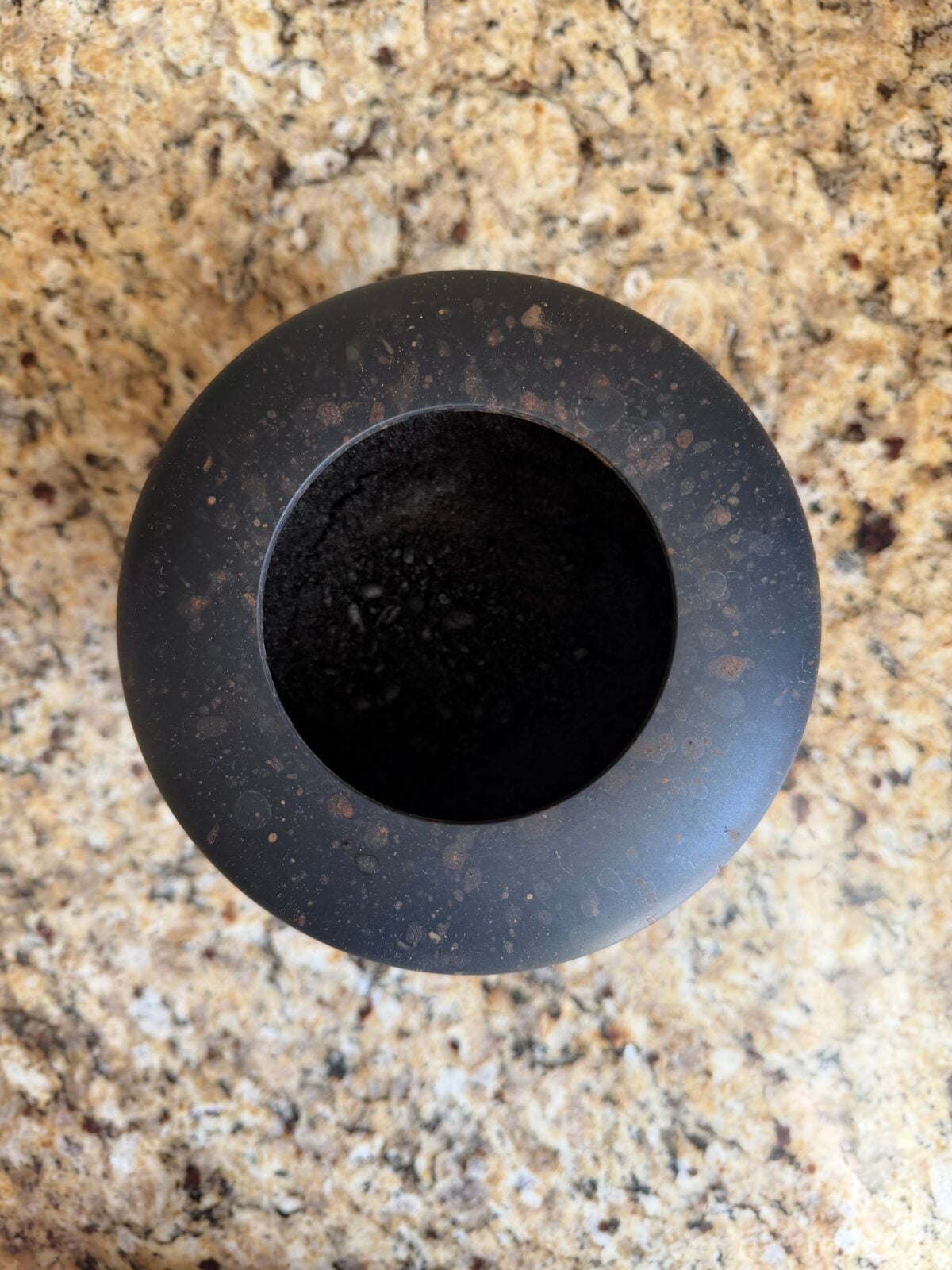 A round, black, stone mortar sits on a speckled beige and brown countertop, viewed from above. The mortar appears empty, and its surface shows some mottled spots.