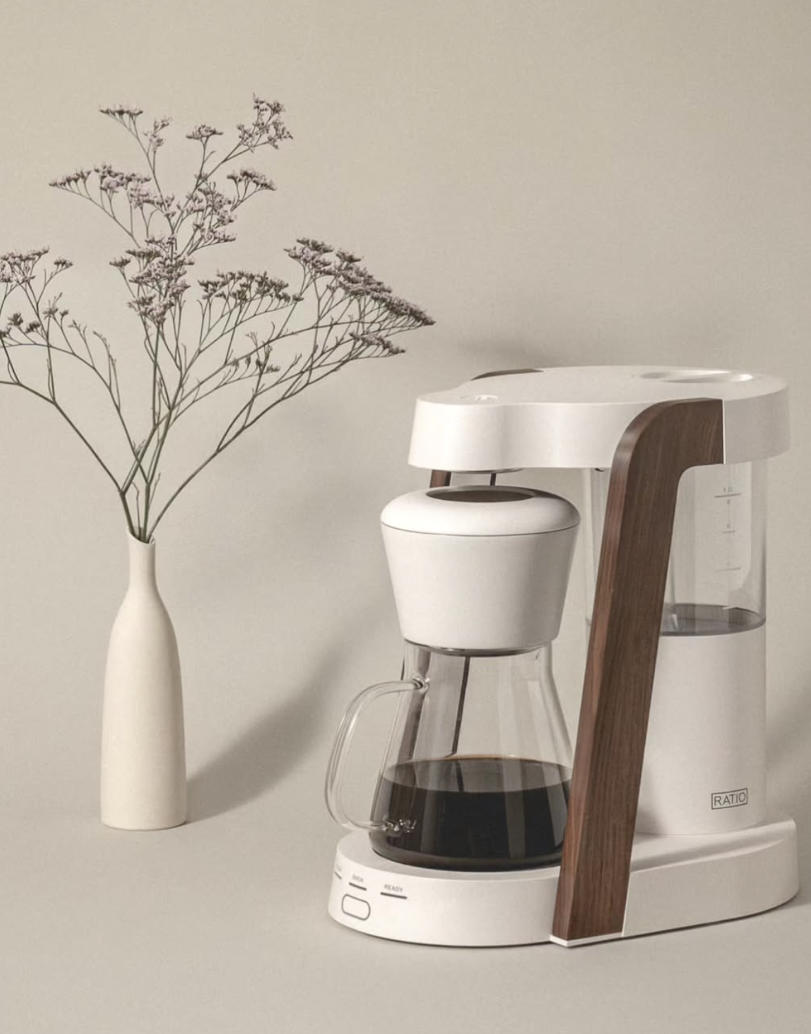 A white and wood-accented drip coffee maker with a glass carafe sits on a beige surface next to a tall white vase holding delicate, sparse branches with small purple flowers.