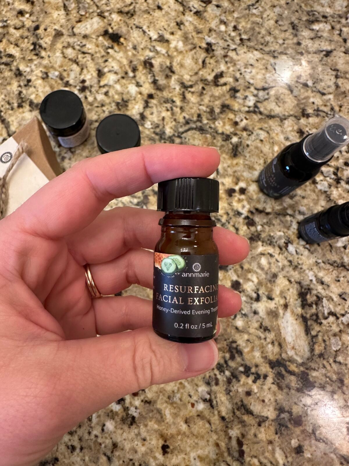 A hand holds a small brown bottle labeled Resurfacing Facial Exfoliant, Honey-Derived Evening Treatment, 0.2 fl oz / 5 mL by Annmarie. Other bottles and items are visible on a granite countertop.