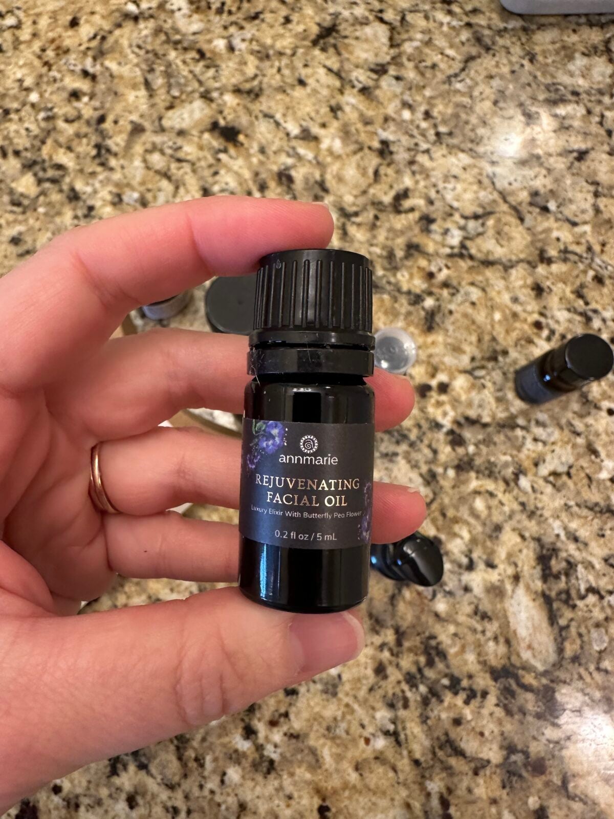 A hand is holding a small dark bottle labeled annmarie Rejuvenating Facial Oil against a granite countertop background. A few other small bottles and caps are visible on the counter.