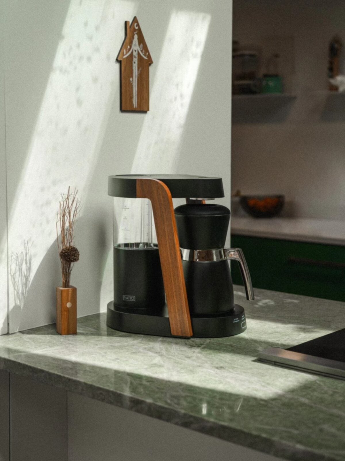 A modern coffee maker with wood accents sits on a green marble countertop in a kitchen. Sunlight streams through a window, casting shadows on the wall. A small vase with dried plants is nearby.