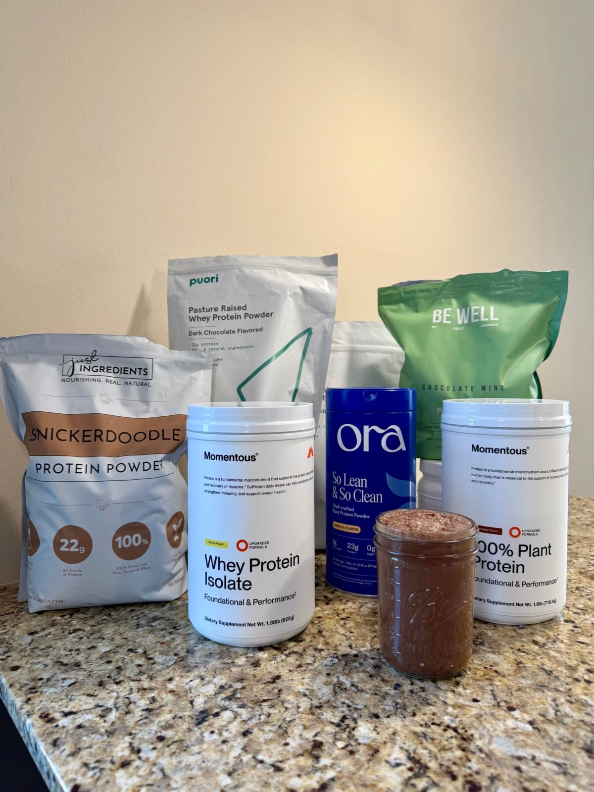 Several protein powder containers and bags from different brands are arranged on a granite countertop, with a glass jar containing a chocolate-colored shake placed in front of them. The background is a plain, light-colored wall.