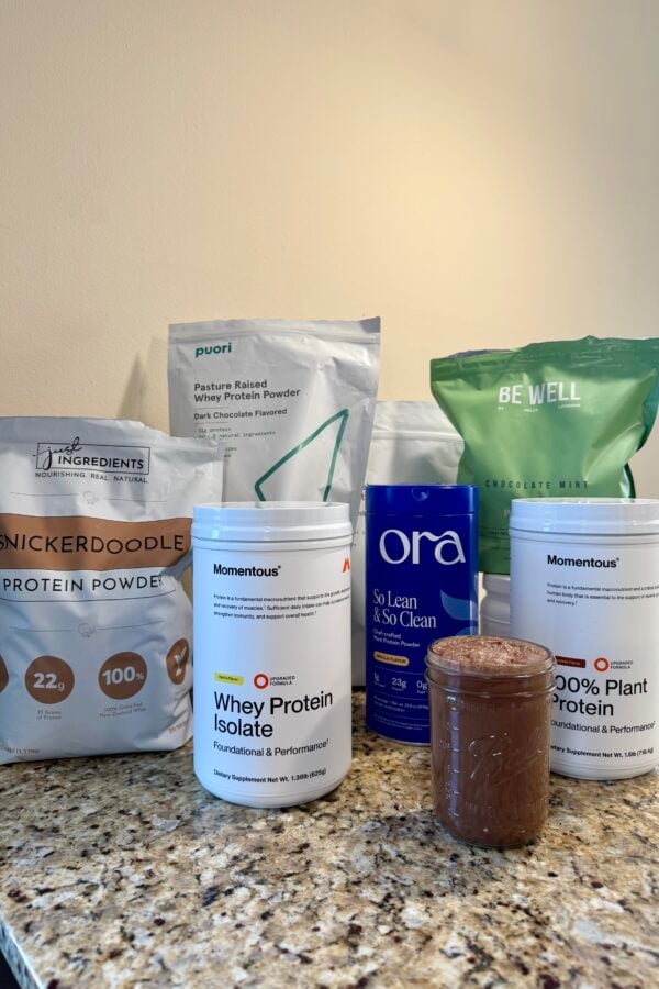 Several protein powder containers and bags from different brands are arranged on a granite countertop, with a glass jar containing a chocolate-colored shake placed in front of them. The background is a plain, light-colored wall.
