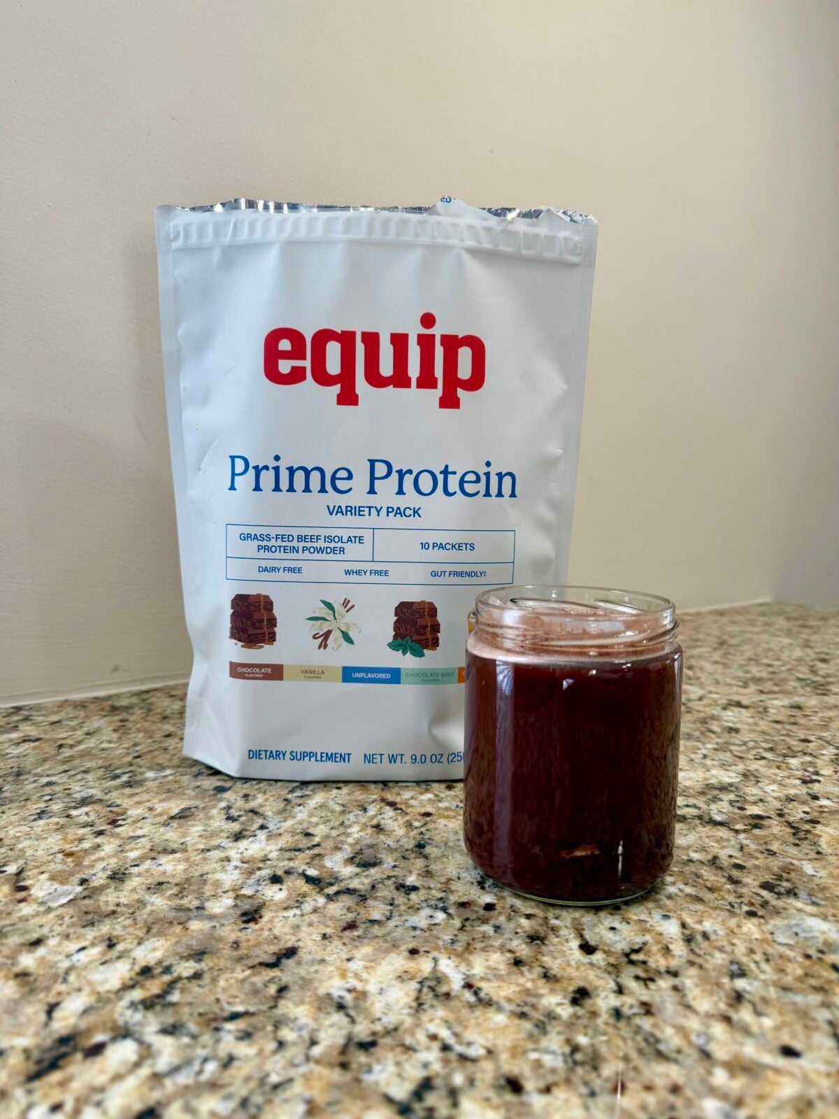 A white bag labeled equip Prime Protein Variety Pack is placed on a granite countertop next to a small glass filled with a dark red liquid.