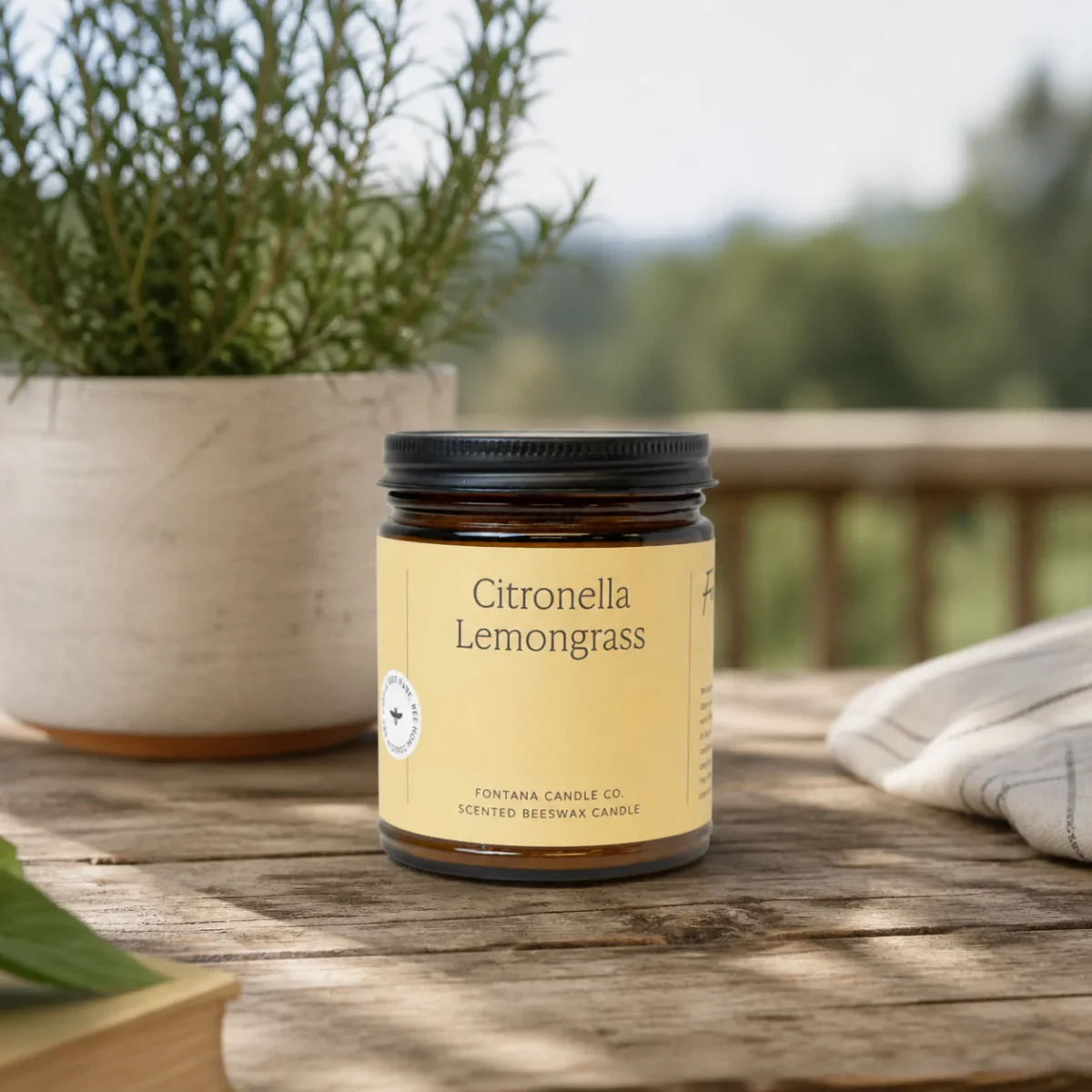 A glass jar labeled Citronella Lemongrass scented beeswax candle sits on a wooden table next to a potted plant, with greenery and a blurred outdoor background.