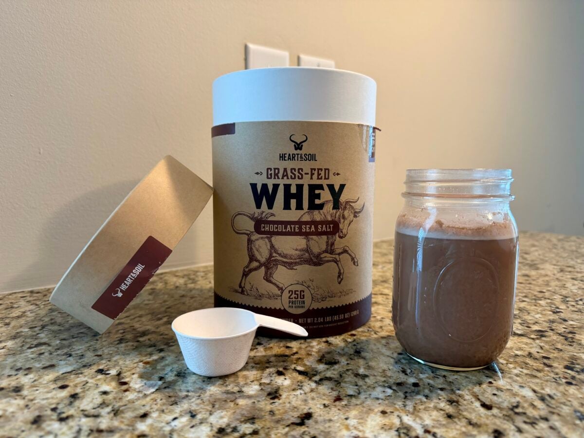 A container of Heart & Sol Grass-Fed Whey protein powder, a white scoop, a lid, and a glass jar filled with chocolate-colored protein shake are on a granite countertop.