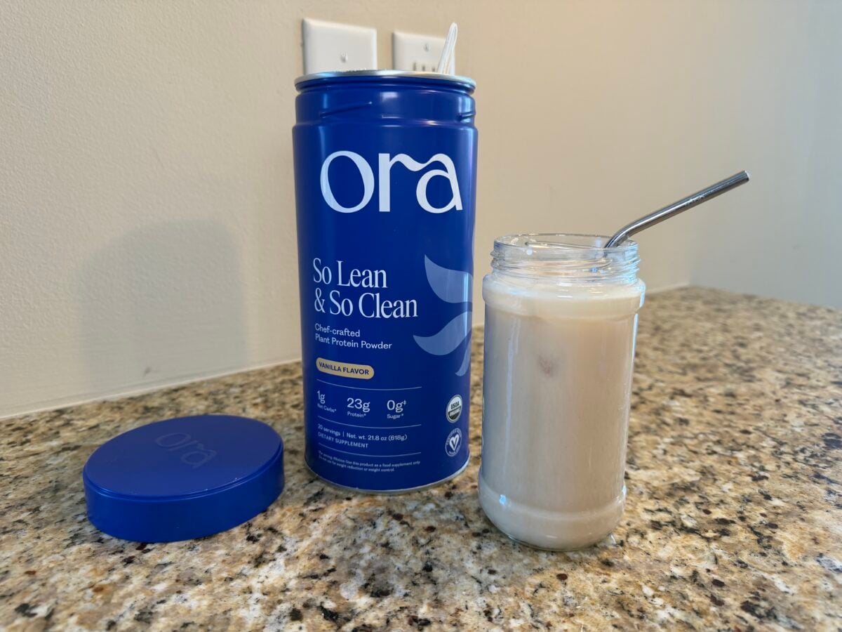 A canister of Ora So Lean & So Clean protein powder stands open on a granite countertop next to its lid. Beside it is a glass jar filled with a creamy drink and a metal straw.