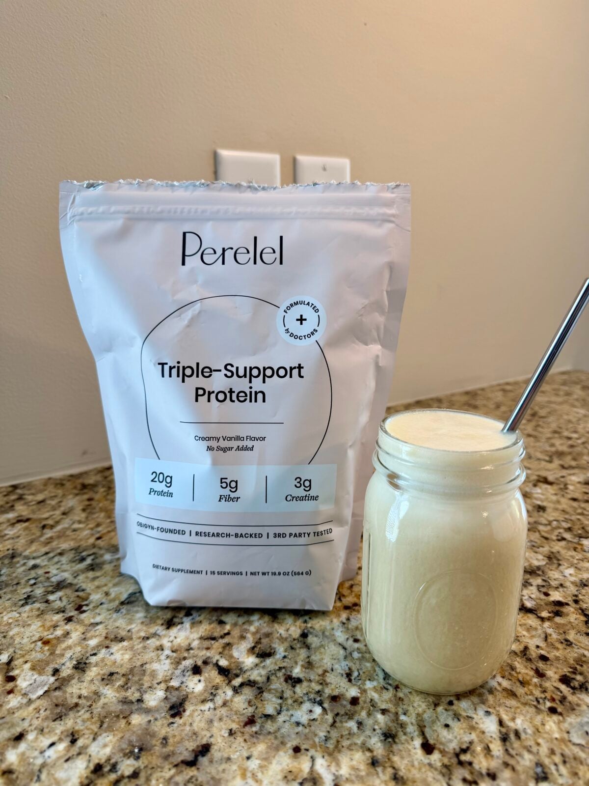 A bag labeled &ldquo;Perelel Triple-Support Protein&rdquo; stands on a granite countertop next to a glass jar filled with a light-colored smoothie and a metal straw. The bag highlights protein, fiber, and creatine content.
