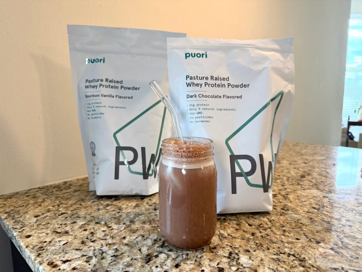 Two bags of Puori pasture raised whey protein powder, one bourbon vanilla flavored and one dark chocolate flavored, are on a granite counter behind a glass filled with a chocolate shake and a clear straw.