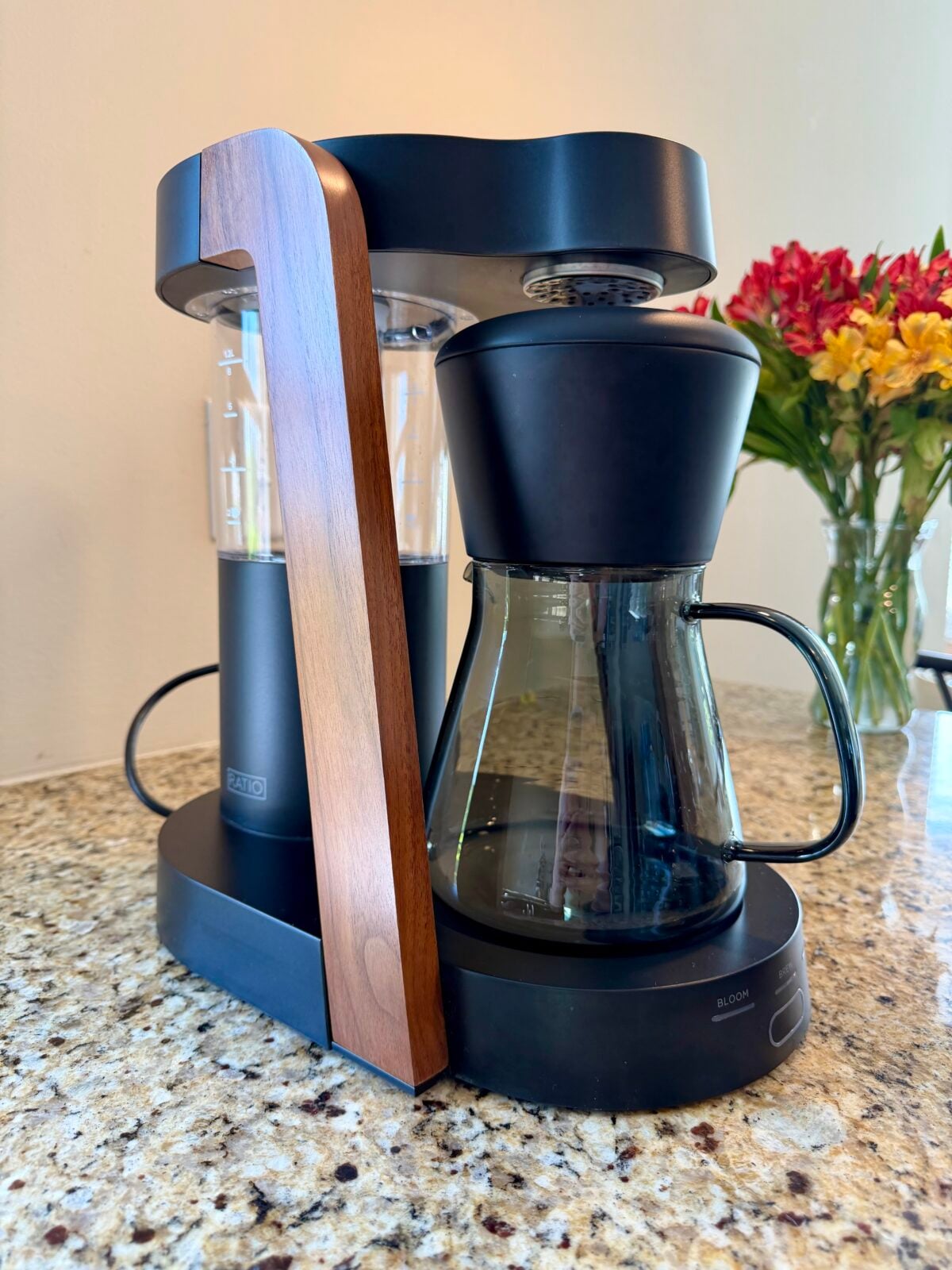 A modern drip coffee maker with a glass carafe and wooden accent sits on a granite countertop. A bouquet of red and yellow flowers in a vase is visible in the background.