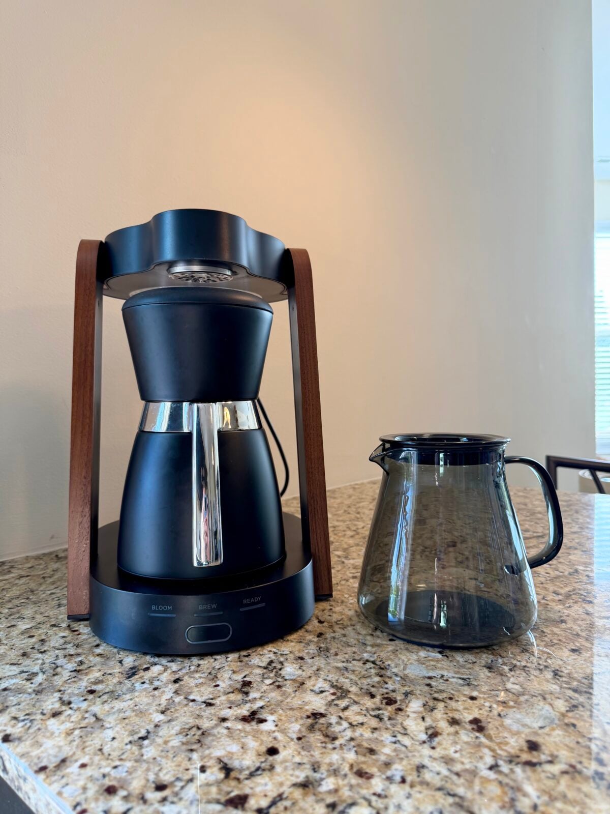 A modern drip coffee maker with a metal carafe stands on a granite countertop next to an empty glass coffee pot. The background features a plain light-colored wall and a window with blinds.