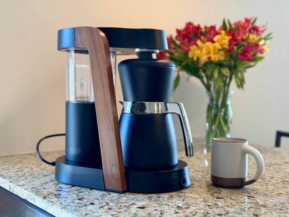 A modern black and wood coffee maker sits on a granite countertop next to a ceramic mug, with a vase of red and yellow flowers in the background.